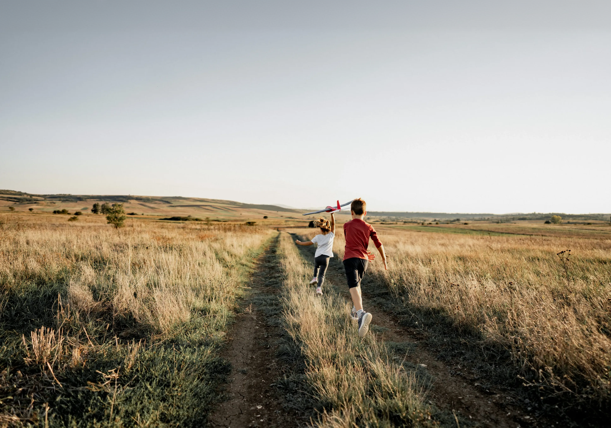 Two young children flying a model airplane down a dirt path through an open field.