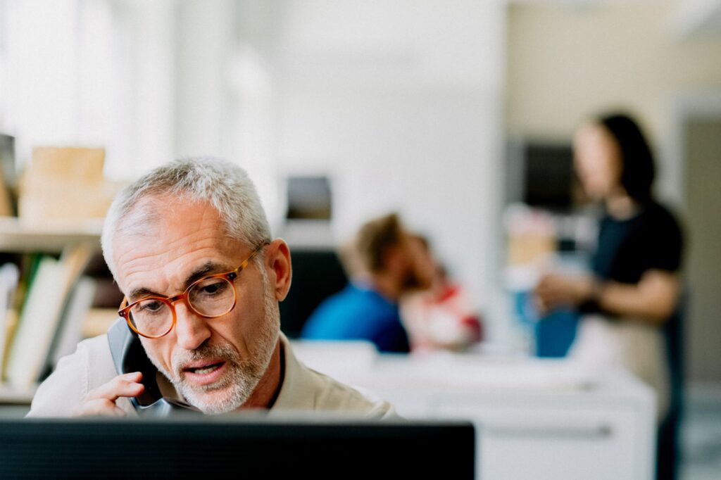 Man at a computer speaking on the phone while other employees chat behind him.